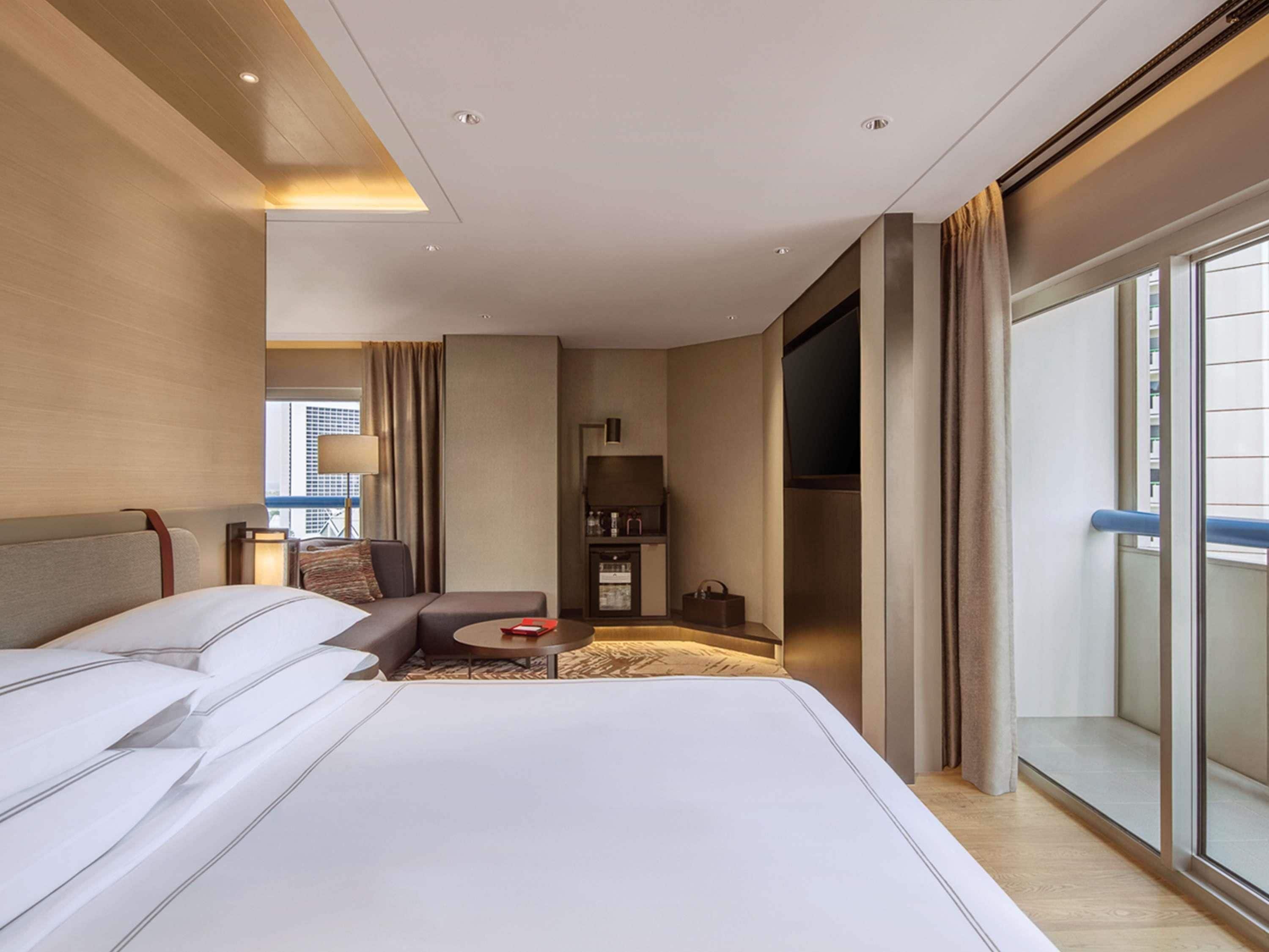A hotel room with a large bed, a seating area with a table, a minibar, and a view of the city.