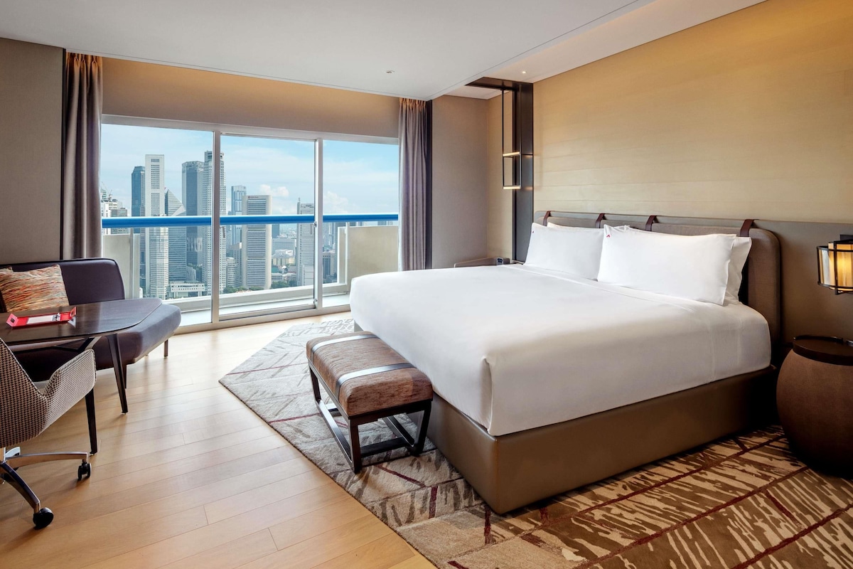 Une chambre d’hôtel avec un grand lit, un bureau, une chaise, une vue sur la ville et un panorama urbain visible par la fenêtre.