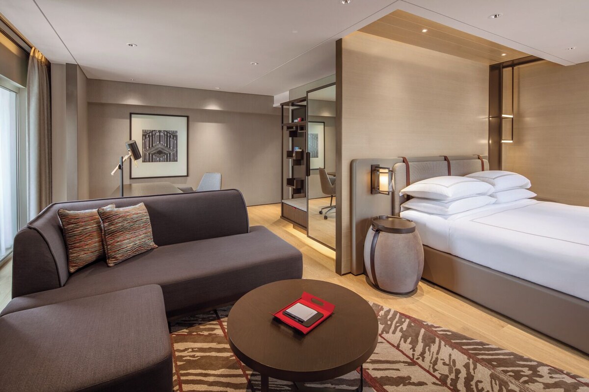 Een moderne hotelkamer met een bank, een rond bijzettafeltje, een bed, een nachtkastje en een groot raam.
