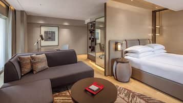 A modern hotel room with a sofa, a round coffee table, a bed, a bedside table, and a large window.