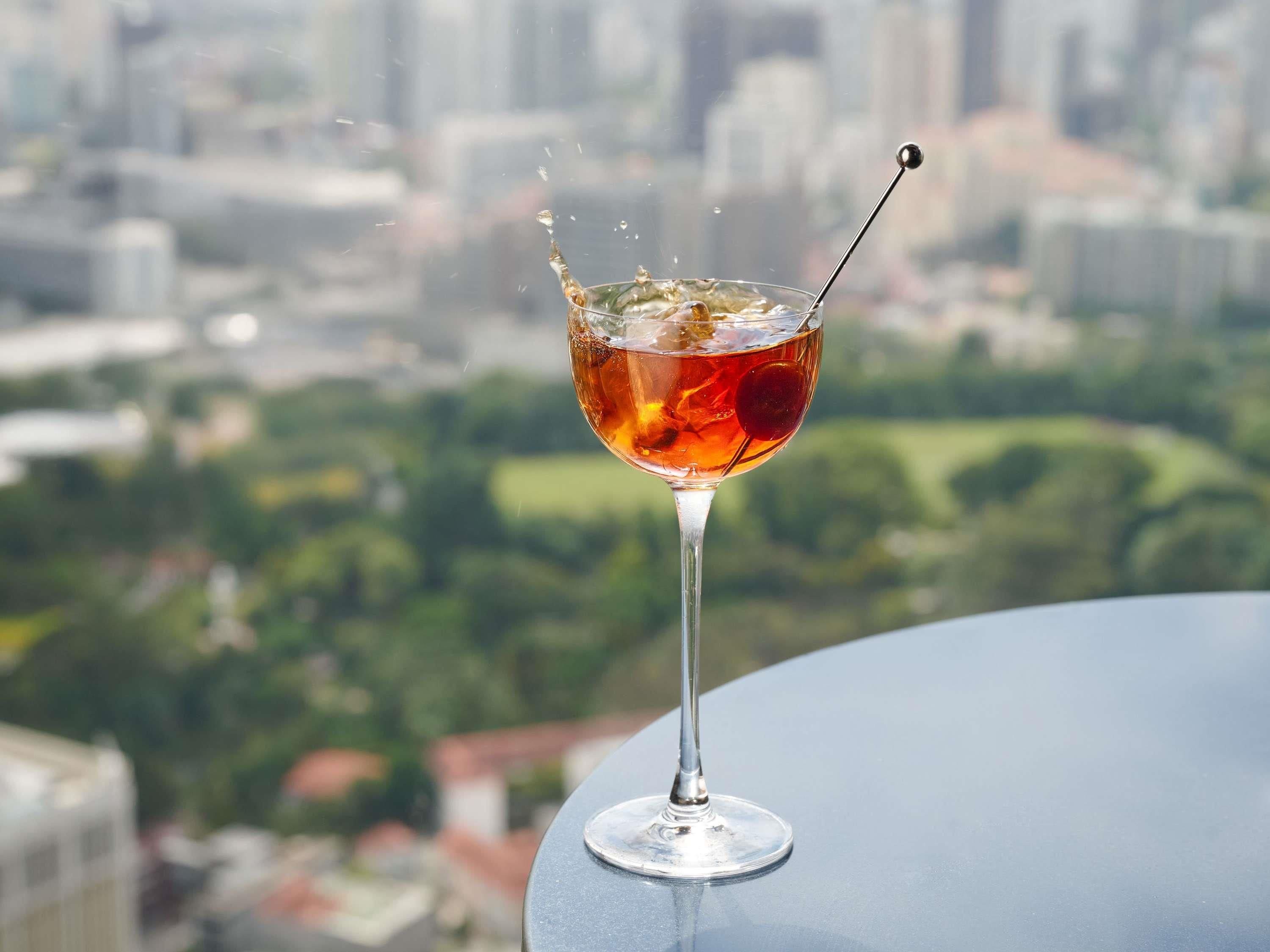 Un verre à cocktail, orné d’une cerise, est posé sur une table devant laquelle s’étend un paysage urbain.