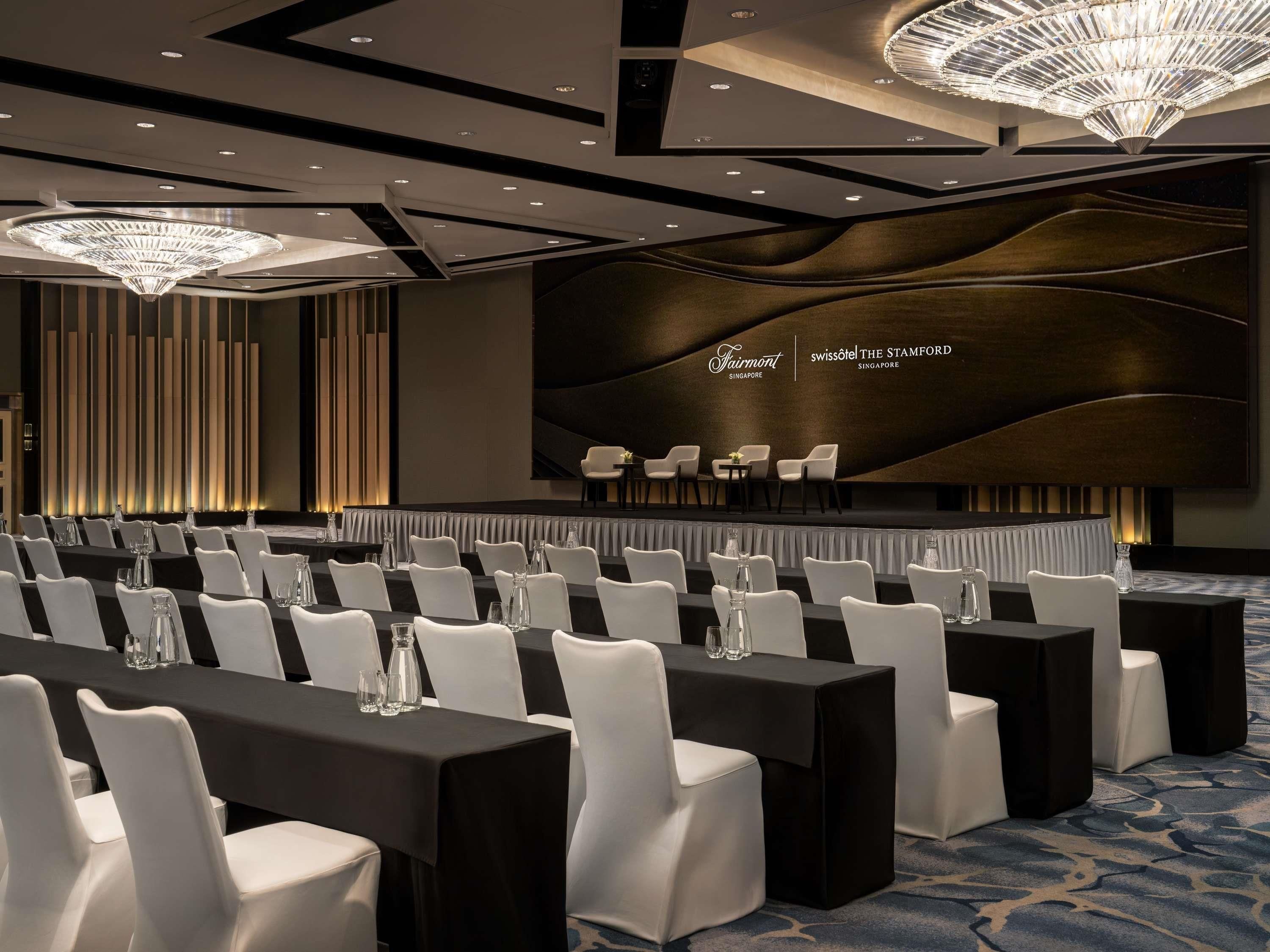A conference room with rows of tables and chairs, a stage, and a large screen displaying the hotel name.