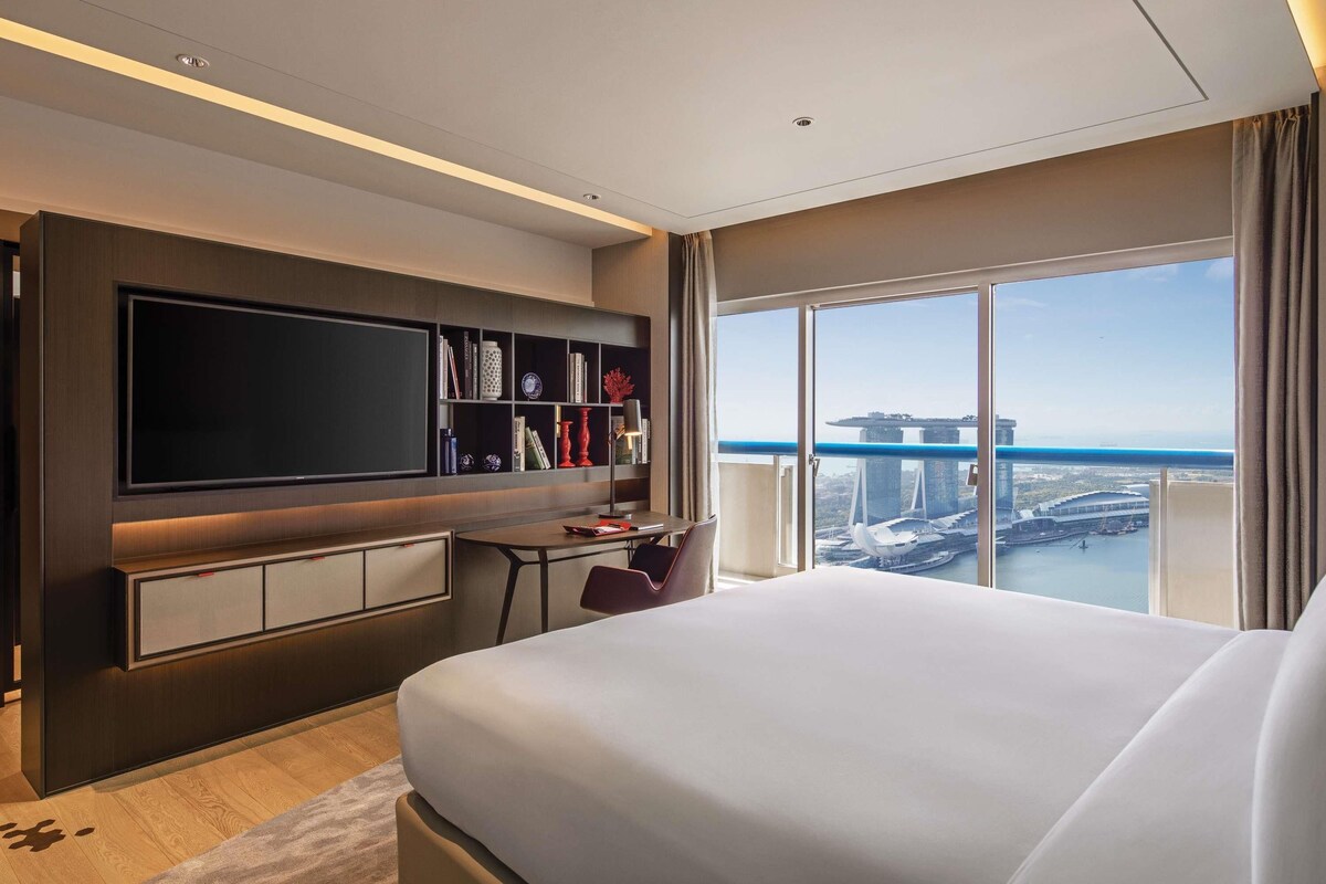 Een moderne hotelkamer met een groot bed, een flatscreen televisie, een bureau en een boekenplank. Een groot raam biedt uitzicht op een stadsbeeld.