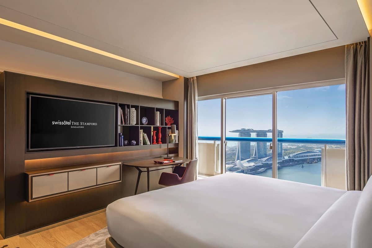 Habitación de hotel con una cama grande, un escritorio con computadora, televisor montado en la pared y vista a la ciudad.