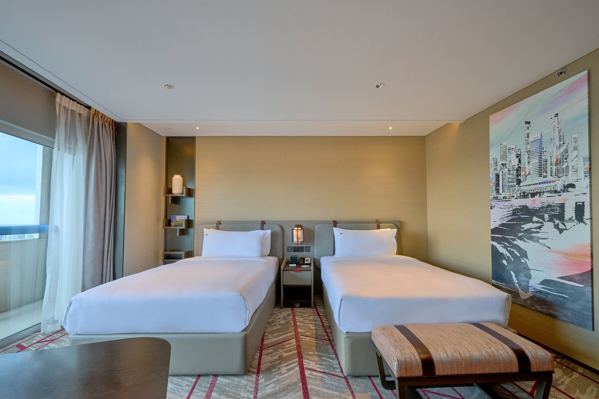 Een hotelkamer met twee bedden, een nachtkastje, een bankje, een groot raam met gordijnen en een stadsgezichtschilderij aan de muur.