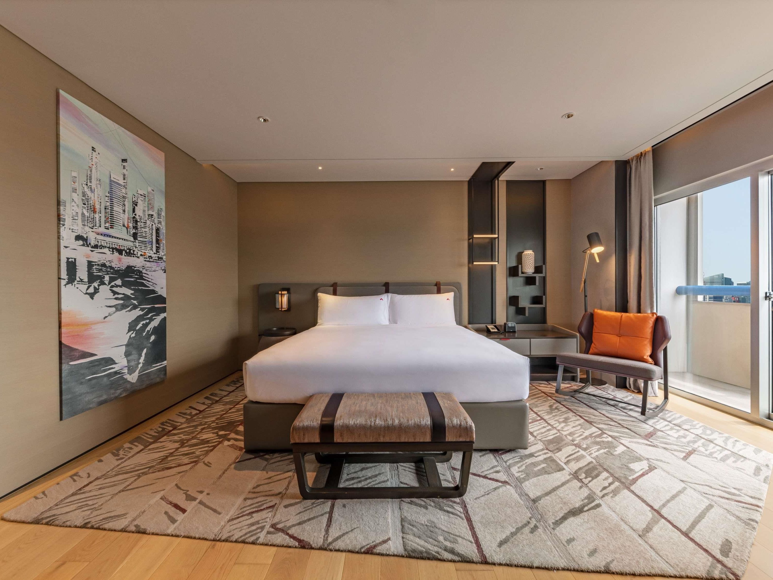 A hotel room with a large bed, a desk, a chair, a balcony with a view, and a painting on the wall.
