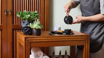 Een houten tafel met een plant in pot, een theepot die in een kopje schenkt, en een dienblad met eten en handdoeken.