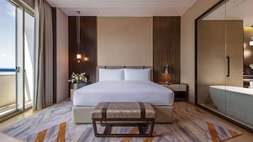 A modern hotel room with a large bed, a bedside table with a lamp, a bench, and a view of the sea through a sliding door.