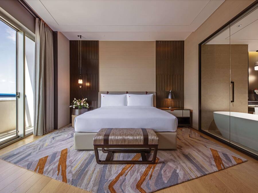 A modern hotel room with a large bed, a bedside table with a lamp, a bench, and a view of the sea through a sliding door.