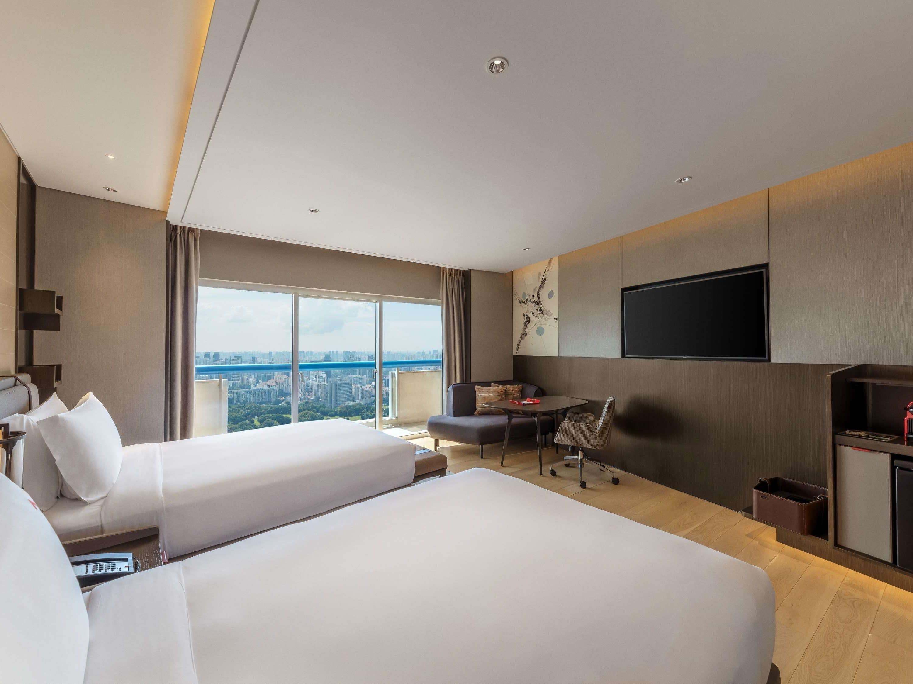 Une chambre d’hôtel avec deux lits, un bureau, une chaise, une télévision et une grande fenêtre donnant sur la ville.