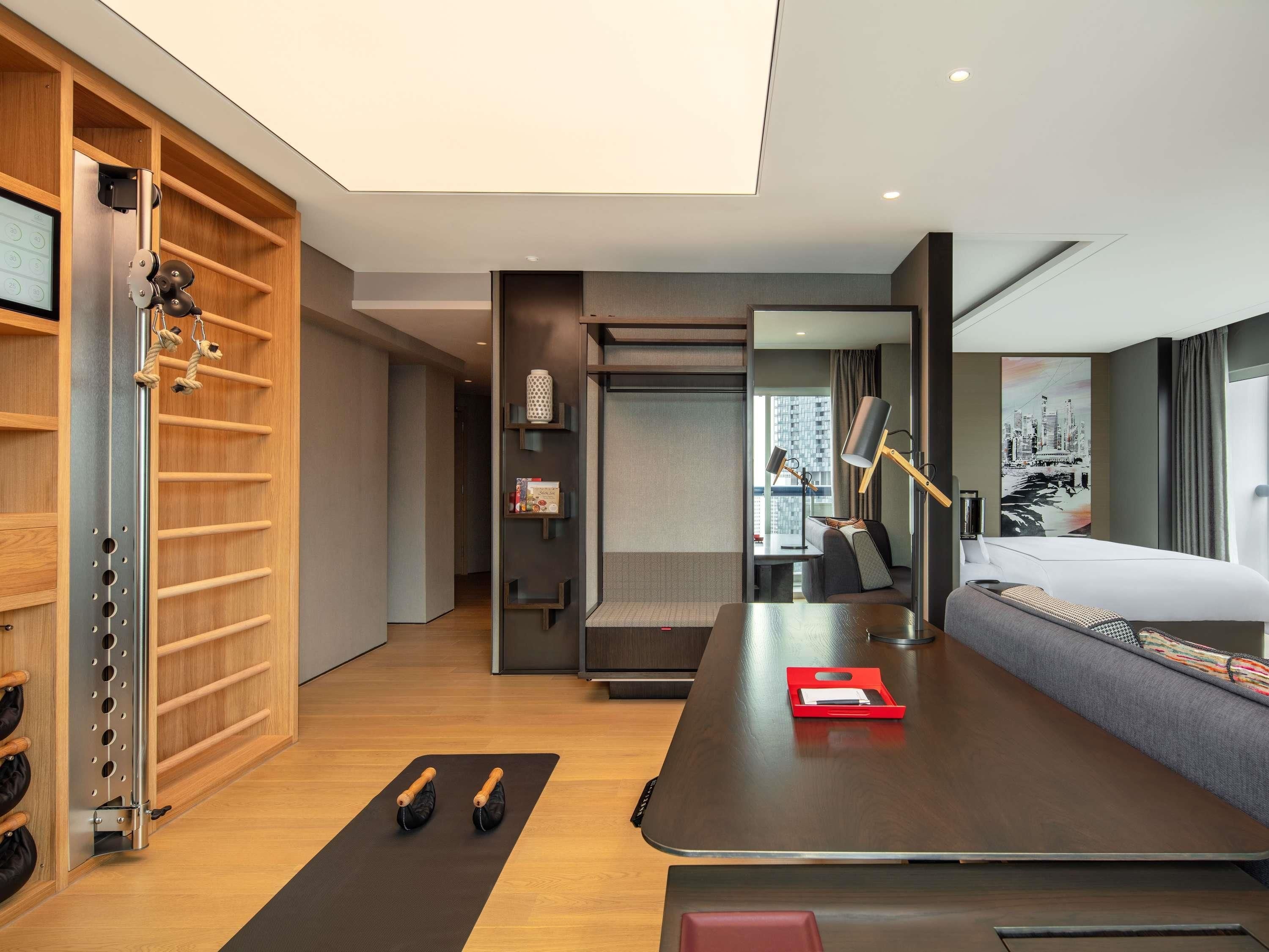 A modern hotel room with a bed, exercise equipment, a desk, and a view of the city.