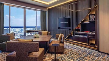 A modern hotel room with a large window, a flat-screen TV, a sofa set, a coffee table, and a patterned carpet.
