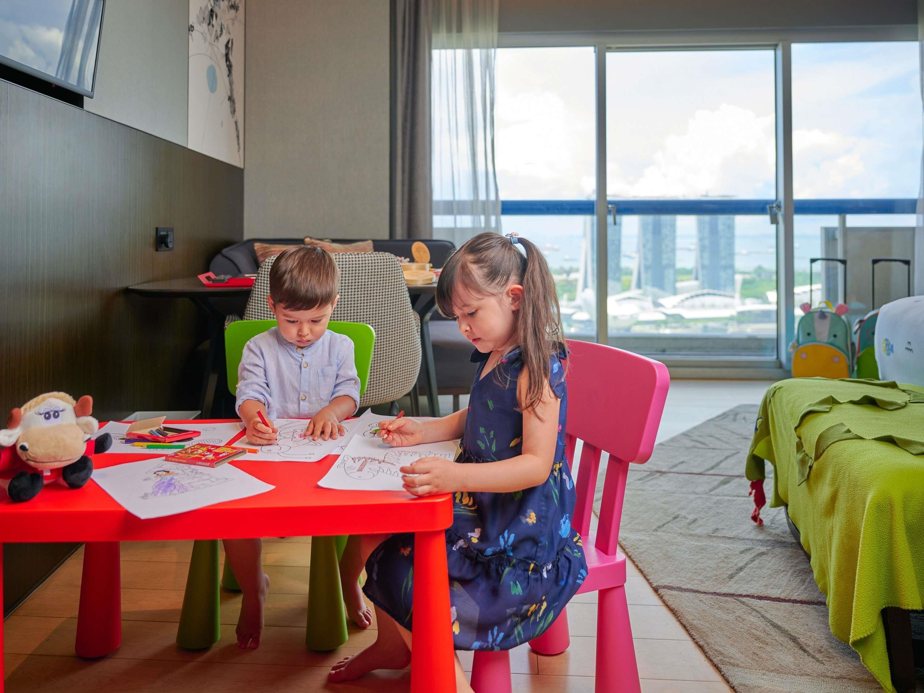 Dos niños dibujando en una mesa roja, con vista a la ciudad.