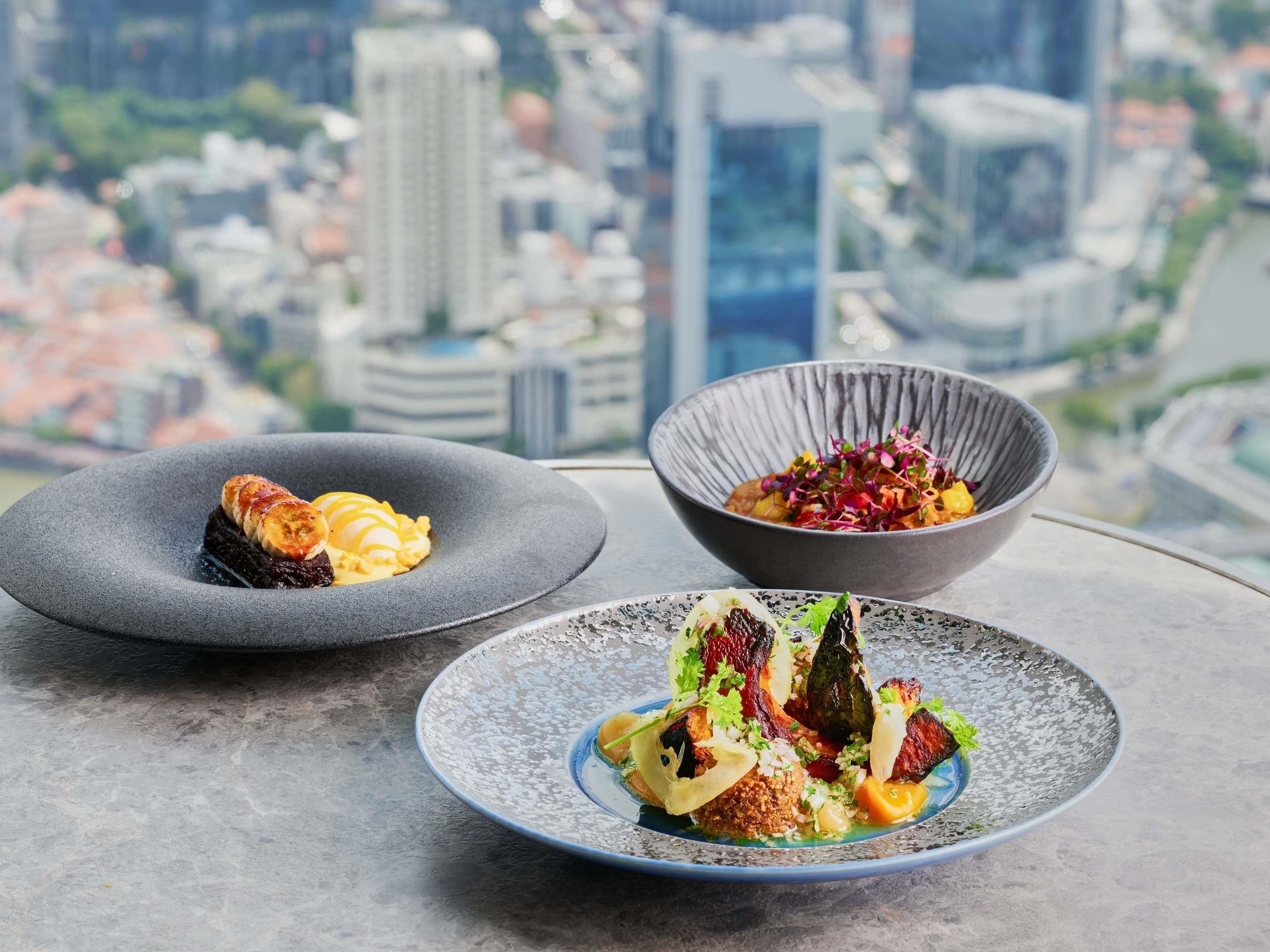 Three dishes on a table with a cityscape view in the background.