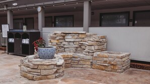 Una jardinera de piedra con un jarrón azul y flores rojas, un letrero que indica una zona infantil, y un área de estar exterior cubierta con suelo de concreto.