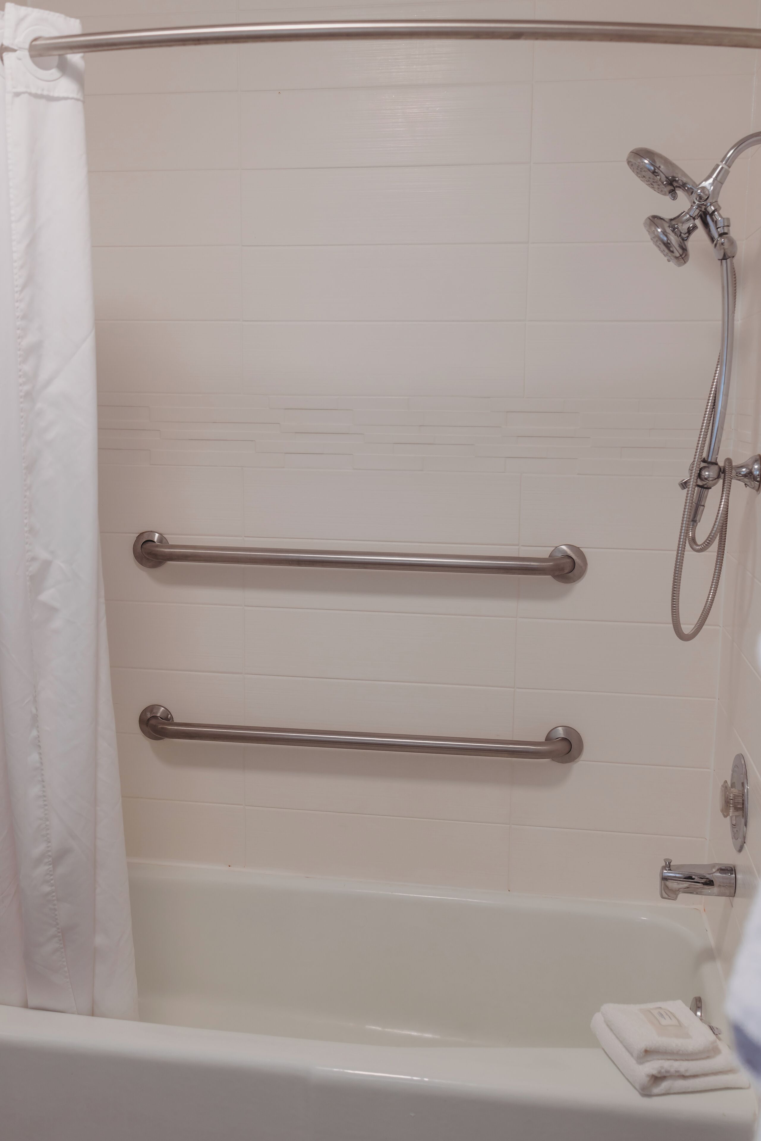Une baignoire blanche équipée de deux barres d’appui métalliques, d’un pommeau de douche et d’une serviette posée sur le bord.