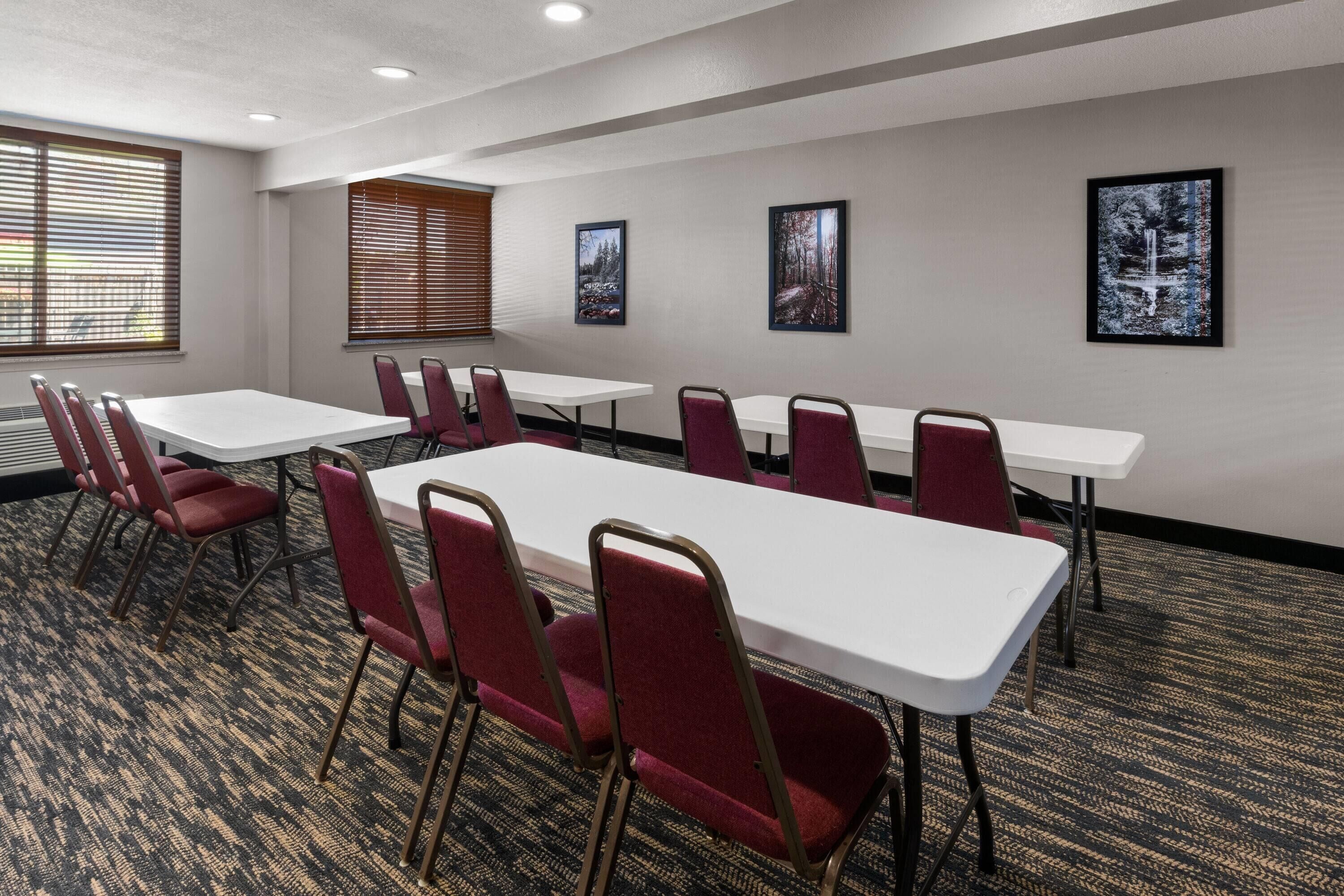 A conference room with white tables and red chairs, large windows with blinds, and framed pictures on the walls.