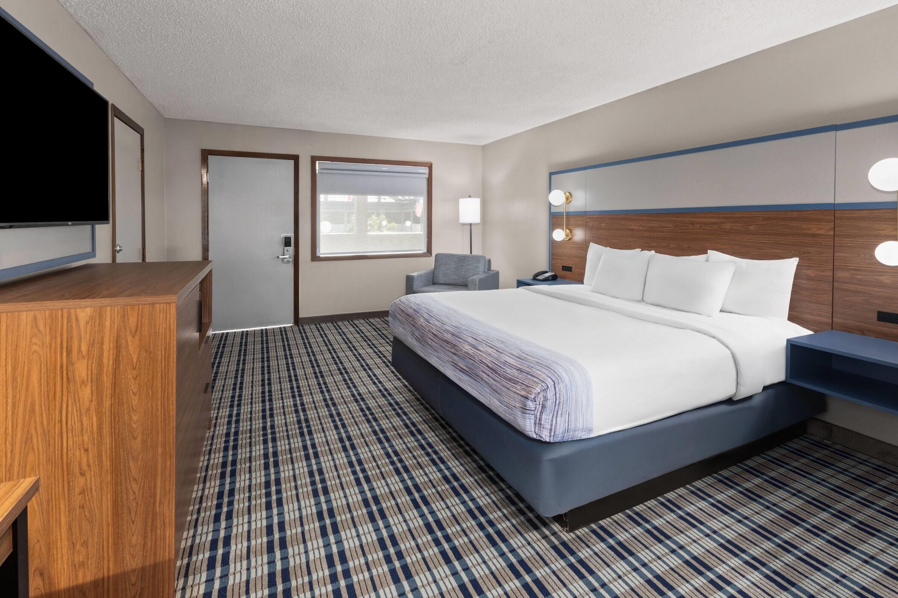 A hotel room with a large bed, a desk, a television, a window with blinds, and a patterned carpet.
