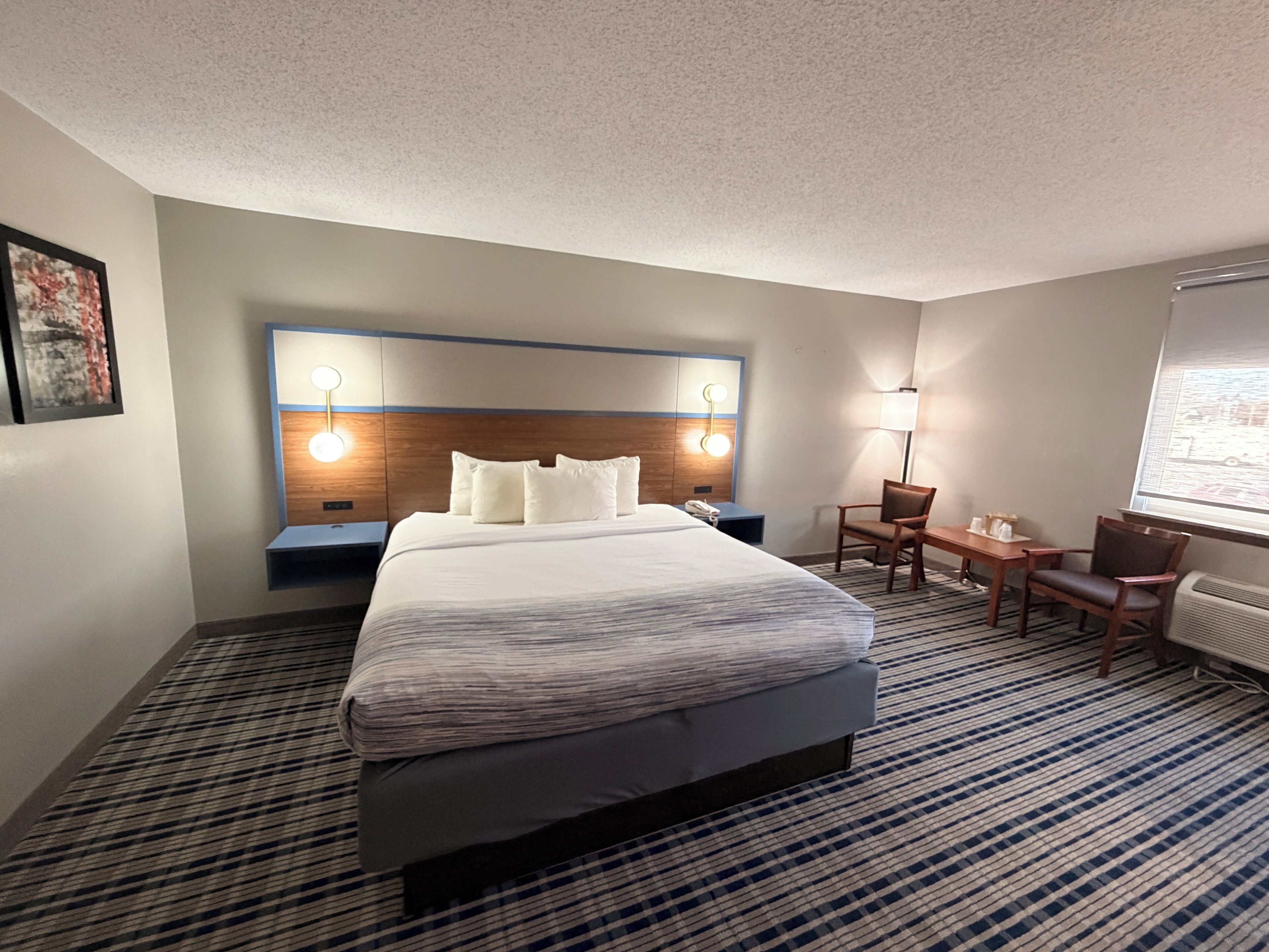 A hotel room with a large bed, bedside tables, a desk with chairs, a lamp, and a window.