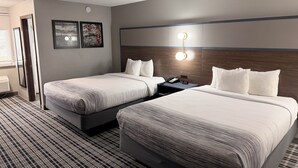 A hotel room with two beds, a nightstand with a phone and clock, a wall-mounted light fixture, and a window with blinds.