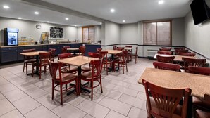 A hotel dining area with multiple tables and chairs, a kitchen in the background, and a clock on the wall.
