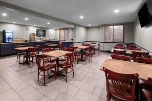 A hotel dining area with multiple tables and chairs, a kitchen in the background, and a clock on the wall.