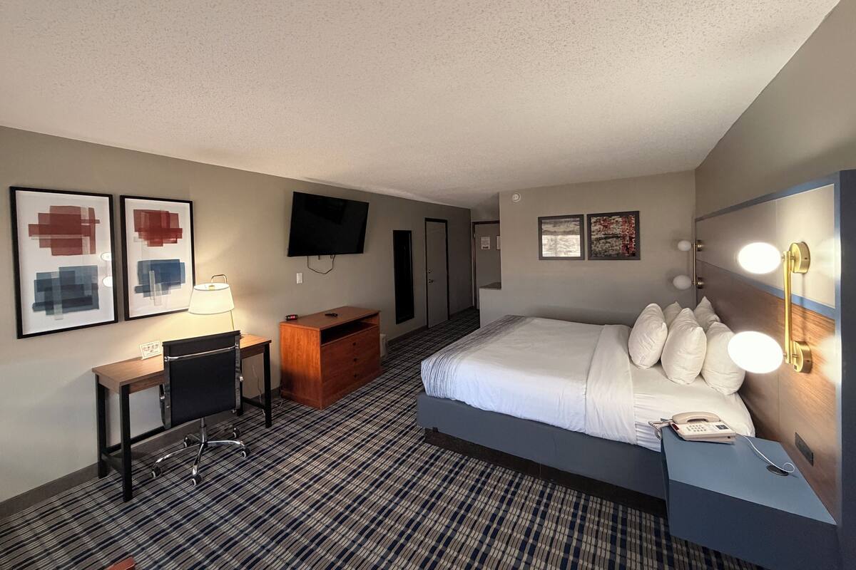 A hotel room with a bed, desk, chair, TV, and artwork on the walls.