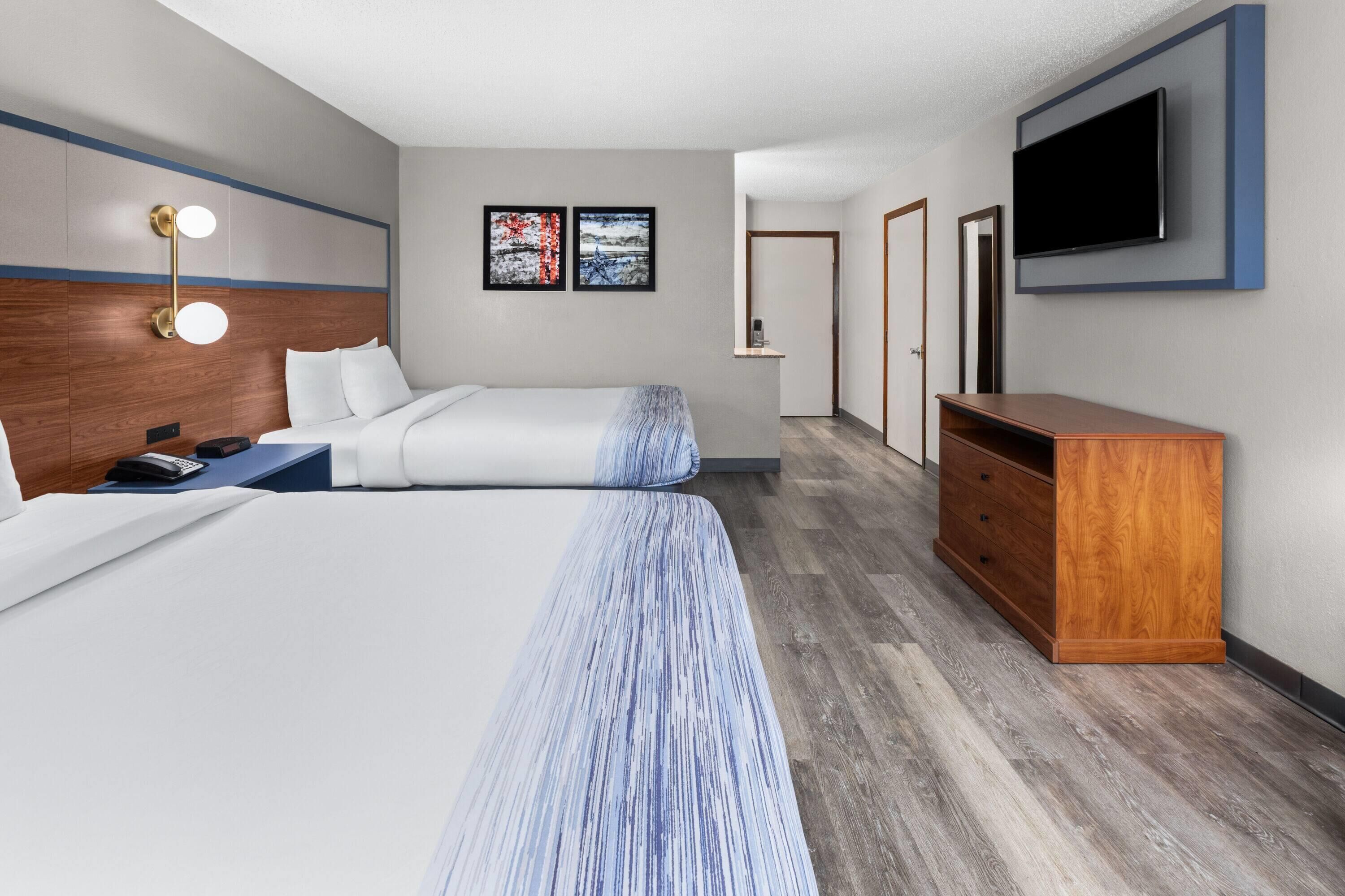 Een hotelkamer met een bed, nachtkastje, lamp, kunstwerk, televisie en een houten dressoir.