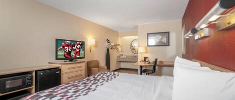 Habitación de hotel con cama, escritorio, silla, televisor, microondas y pequeña cocina.