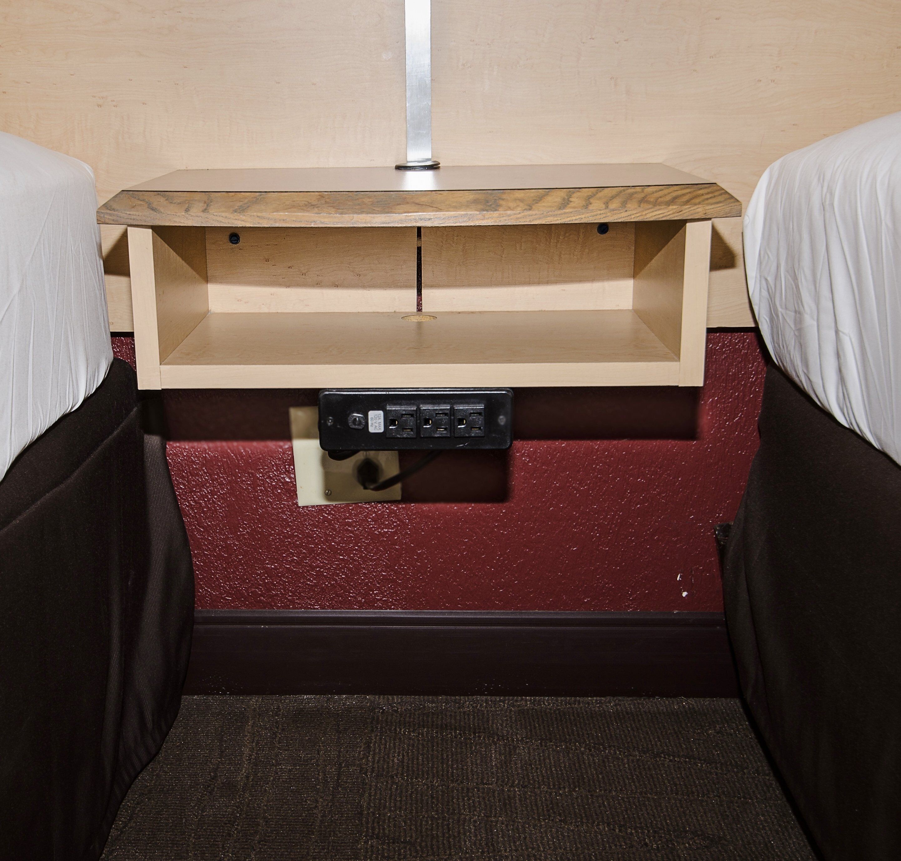 Een ruim bed met daarboven een houten plank boven een stopcontact, een rood structuurwand en een donkere vloer.