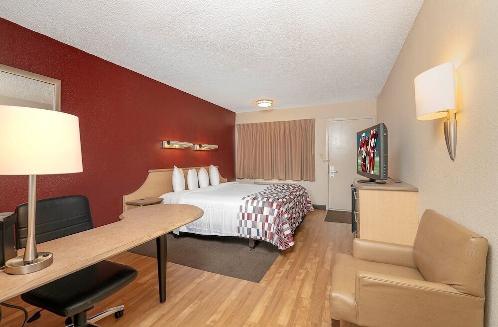 A hotel room with a bed, desk, chair, lamp, TV, and a painting on the wall.