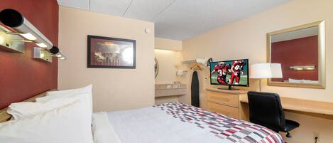 A hotel room with a bed, a desk with a television, a mirror, and a wall-mounted lamp.