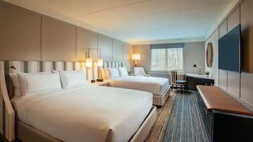 A hotel room with two beds, a desk, a chair, a TV, and a window.