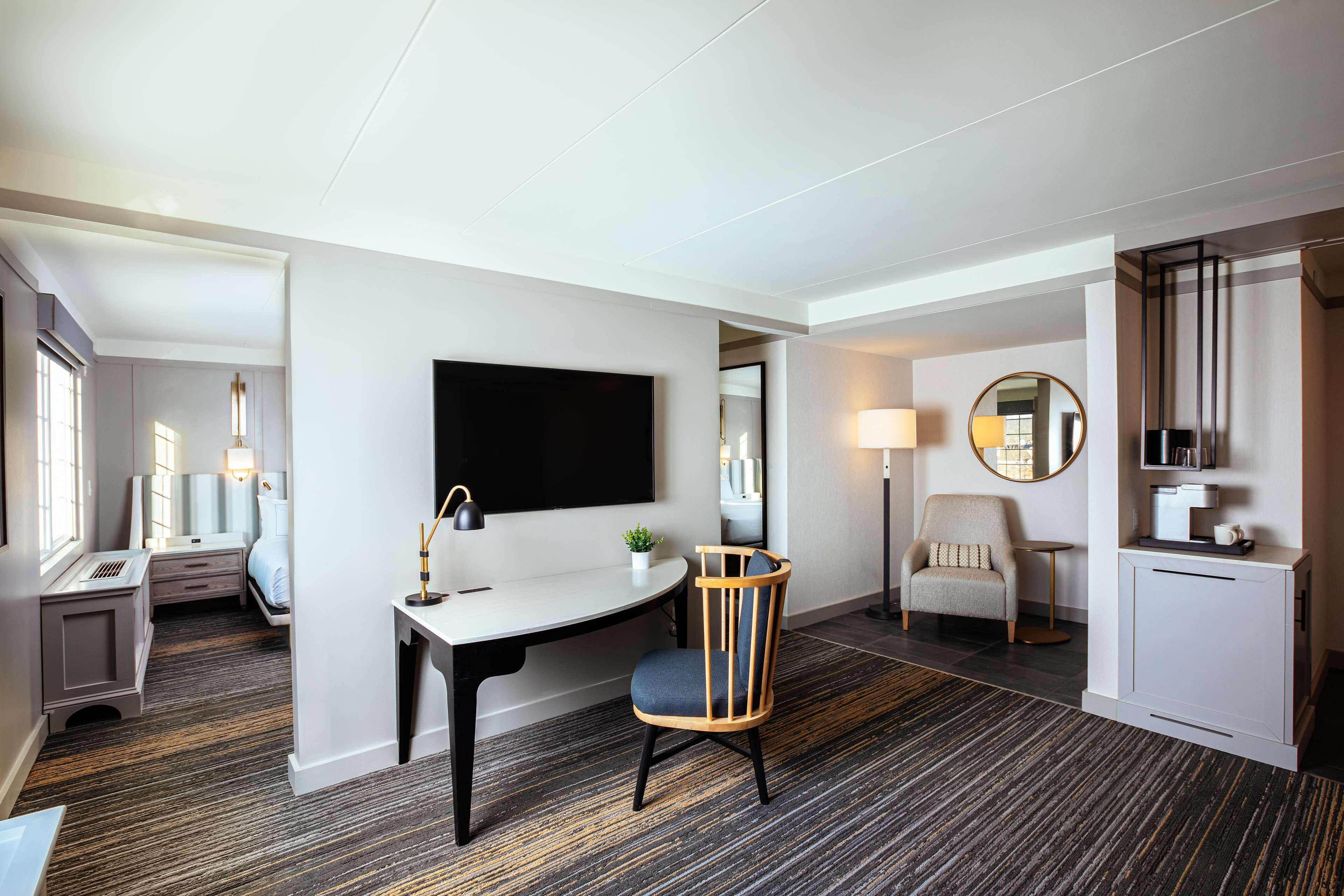 A modern hotel room with a desk, chair, TV, bedside table, lamp, and a small plant.