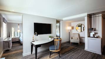 A modern hotel room with a desk, chair, TV, bedside table, lamp, and a small plant.