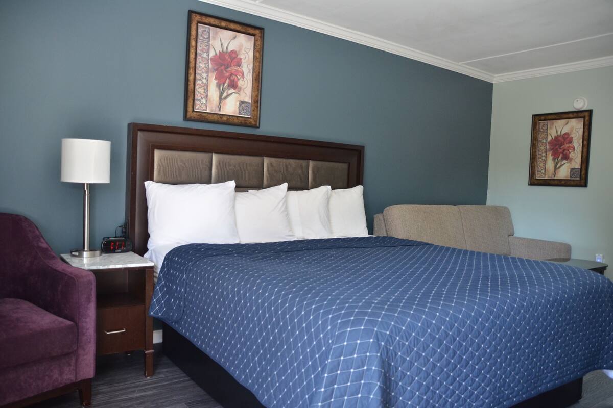 A hotel room with a large bed, a nightstand with a lamp, a chair, and two framed floral paintings on the walls.