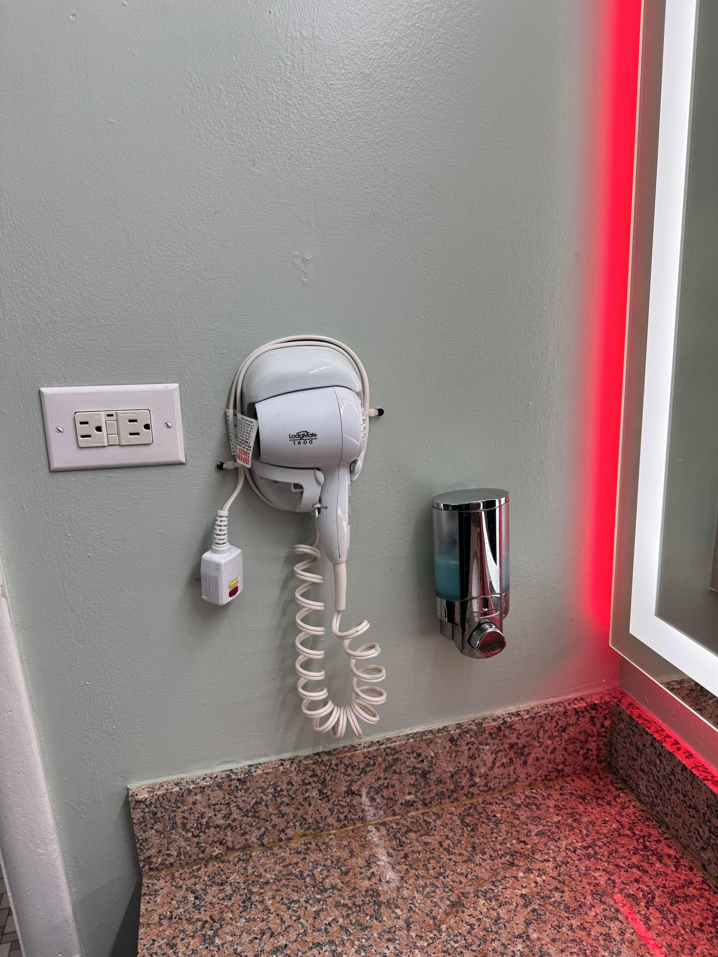 A hair dryer mounted on a wall with a power outlet and a hand sanitizer dispenser nearby.
