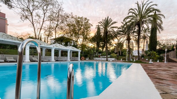 A swimming pool with a white deck and lounge chairs, surrounded by palm trees and a gazebo. - Hotel Exe Guadalete (Jerez de la Frontera)
