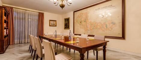 Een vergaderruimte met een grote houten tafel, verschillende stoelen, een kroonluchter, een kaart aan de muur en een boekenkast.