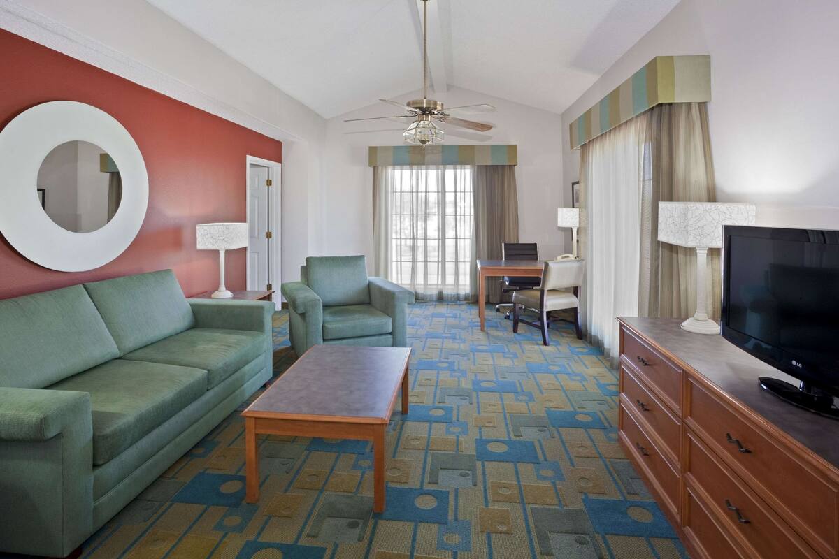Een hotelkamer met een groene bank, een houten salontafel, een televisie, een bureau met een computer en een plafondventilator.