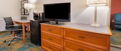 A hotel room with a wooden desk, a flat-screen TV, a lamp, a chair, and a dresser.