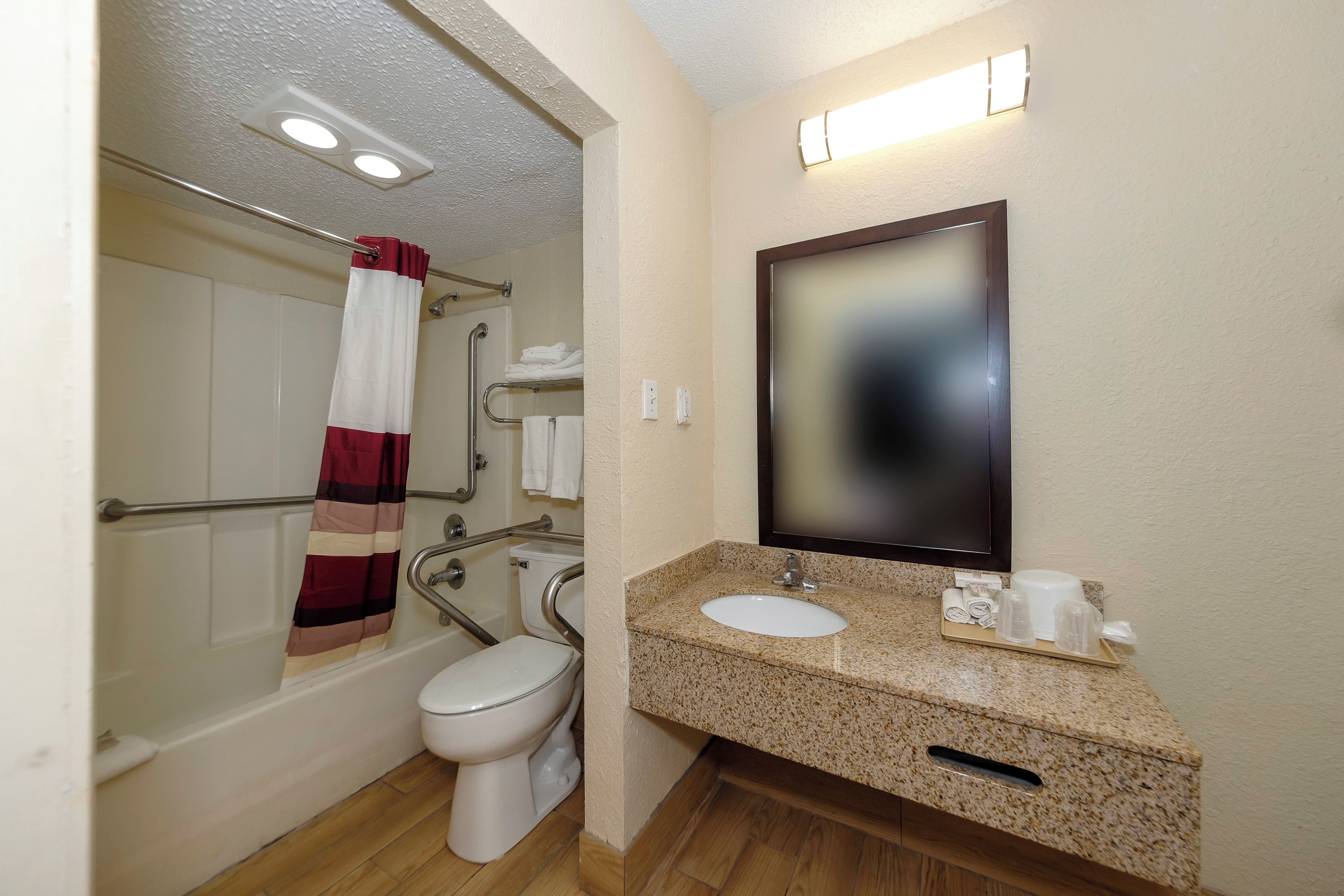 A bathroom with a toilet, grab bars, a shower with a red and white curtain, a sink with a tray of toiletries, and a mirror.