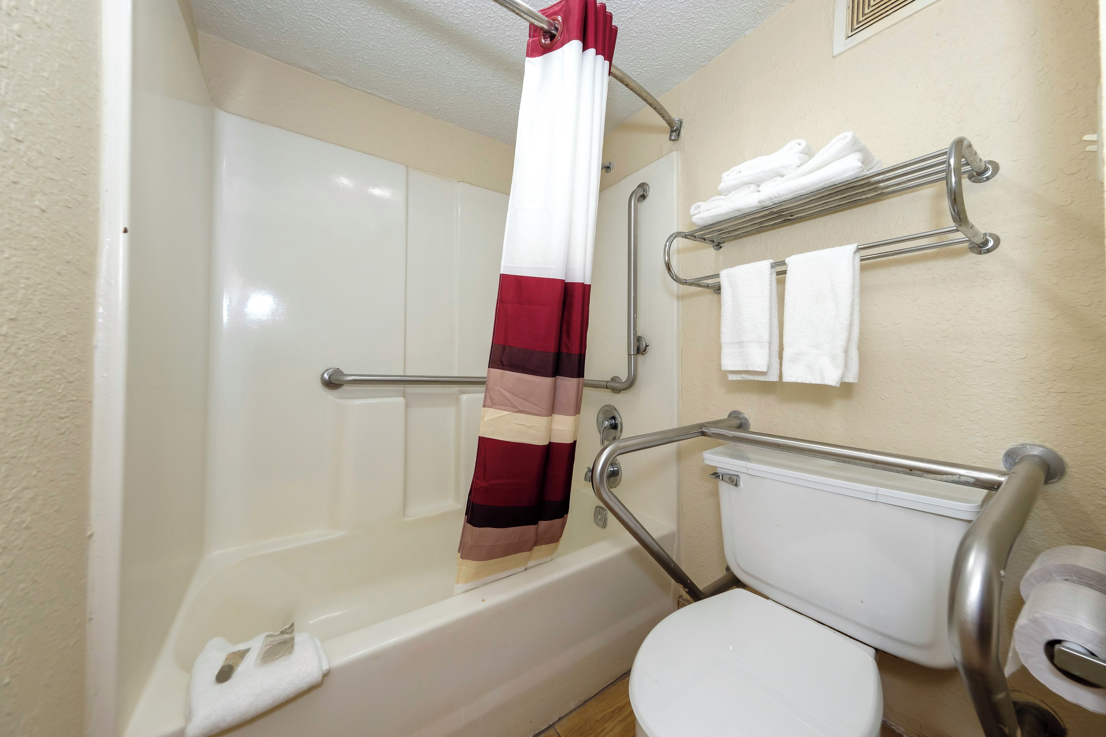 Un baño con un inodoro blanco, una ducha con cortina de baño roja y blanca, y una barra de apoyo. Hay toallas blancas en un perchero.