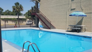Ein rechteckiger Pool mit einer Metallleiter im Vordergrund, einer Treppe, die zu einem Balkon führt, und einem Tisch mit Sonnenschirm im Hintergrund.