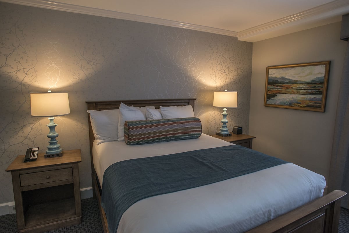 A neatly made bed with a blue blanket, two bedside lamps, a nightstand with a phone, and a painting on the wall.