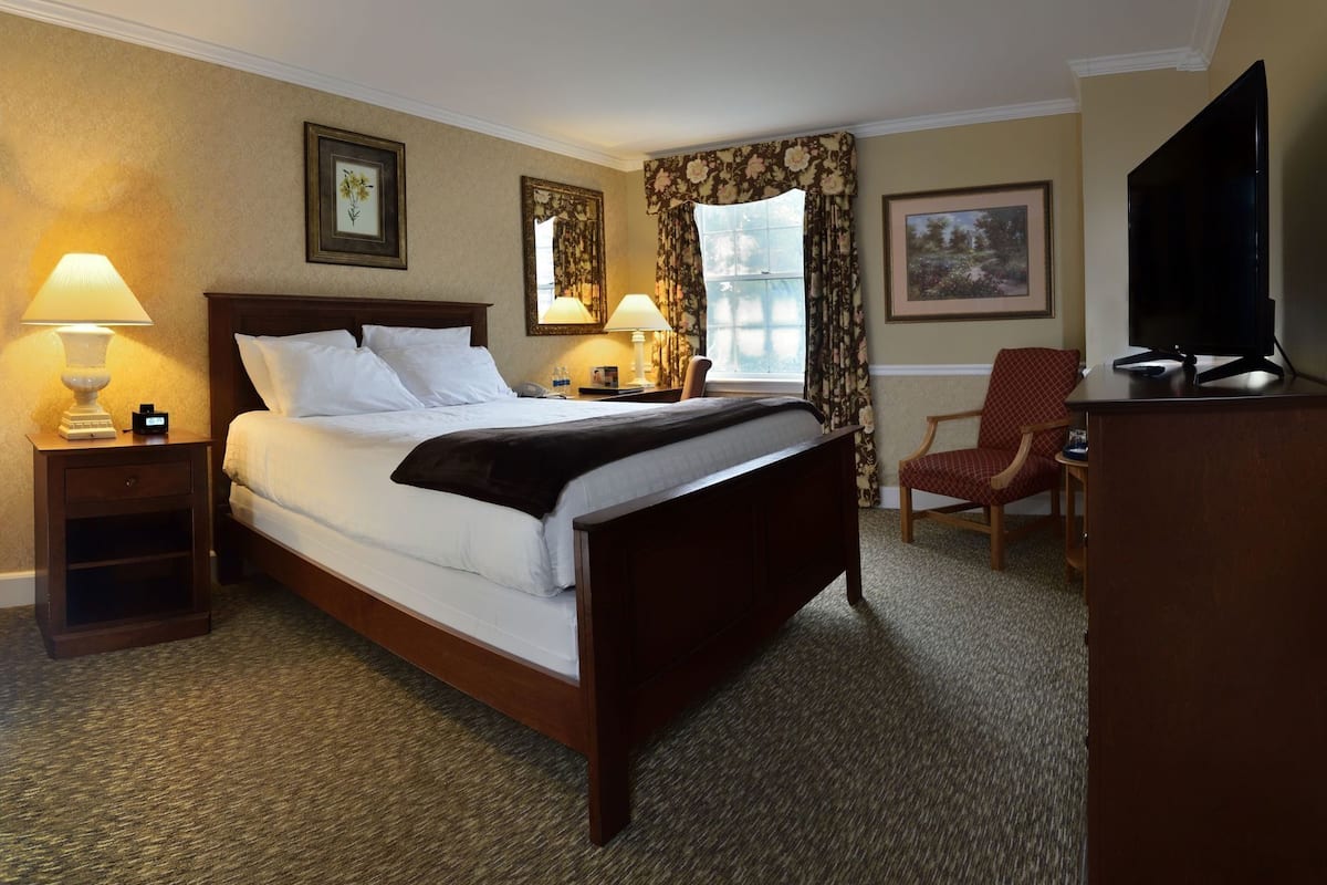 A hotel room with a bed, bedside lamps, a chair, a desk, a television, and framed pictures on the walls.