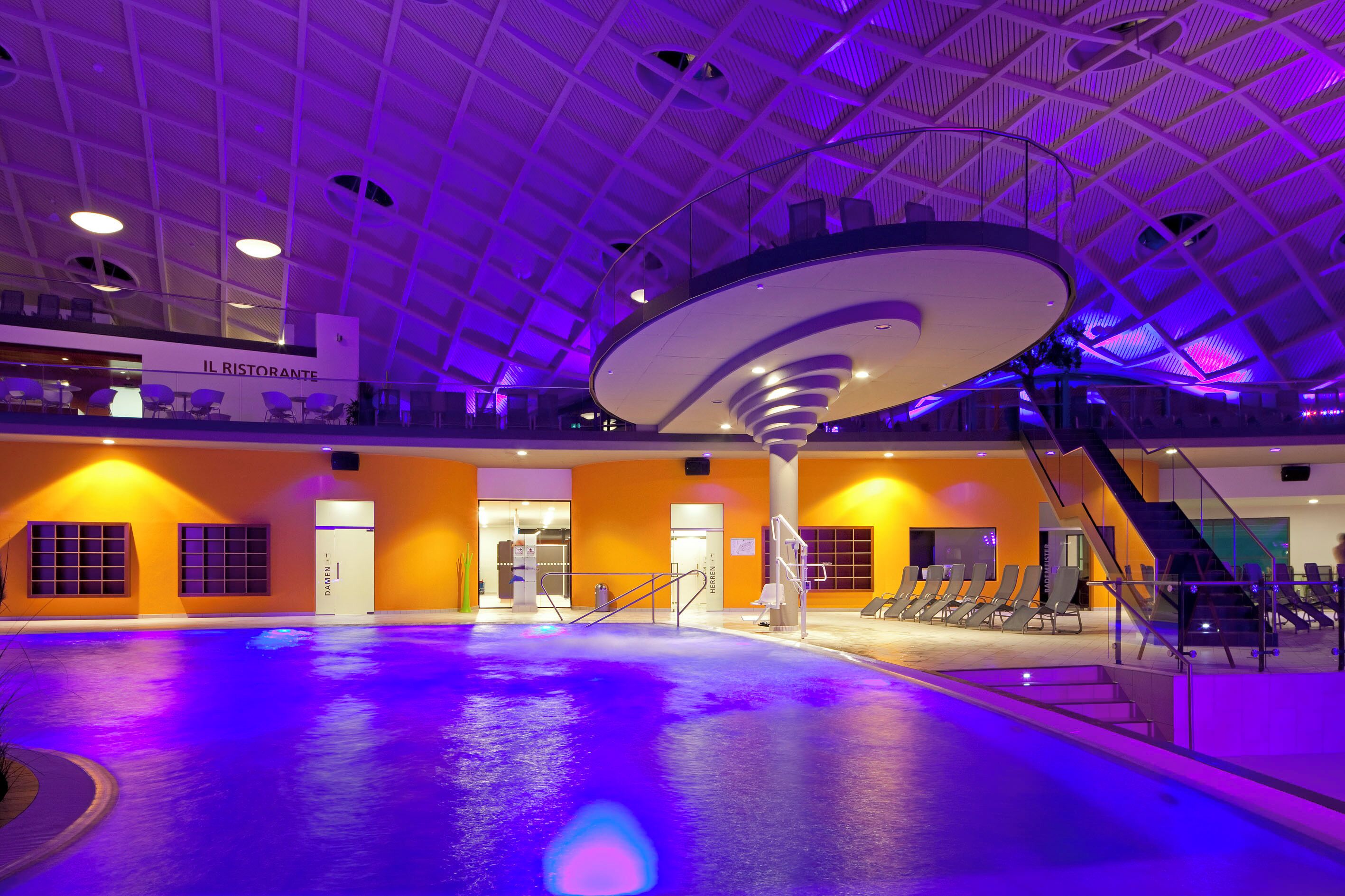 Una piscina interior con un diseño moderno de techo, un cartel que indica 'IL RISTORANTE' y una zona de estar con sillas y una escalera.