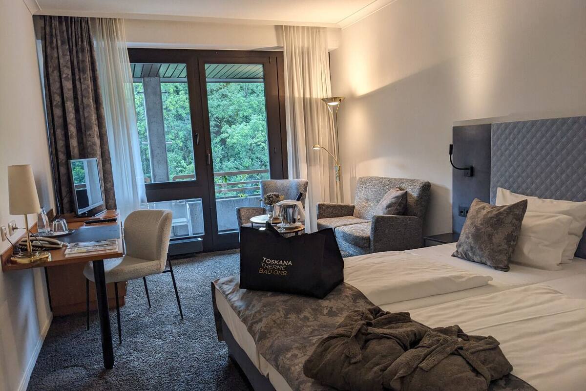 A hotel room with a bed, a desk, a chair, and a sofa. There is a bag on the bed and a lamp on the desk.