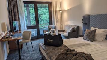 A hotel room with a bed, a desk, a chair, and a sofa. There is a bag on the bed and a lamp on the desk.