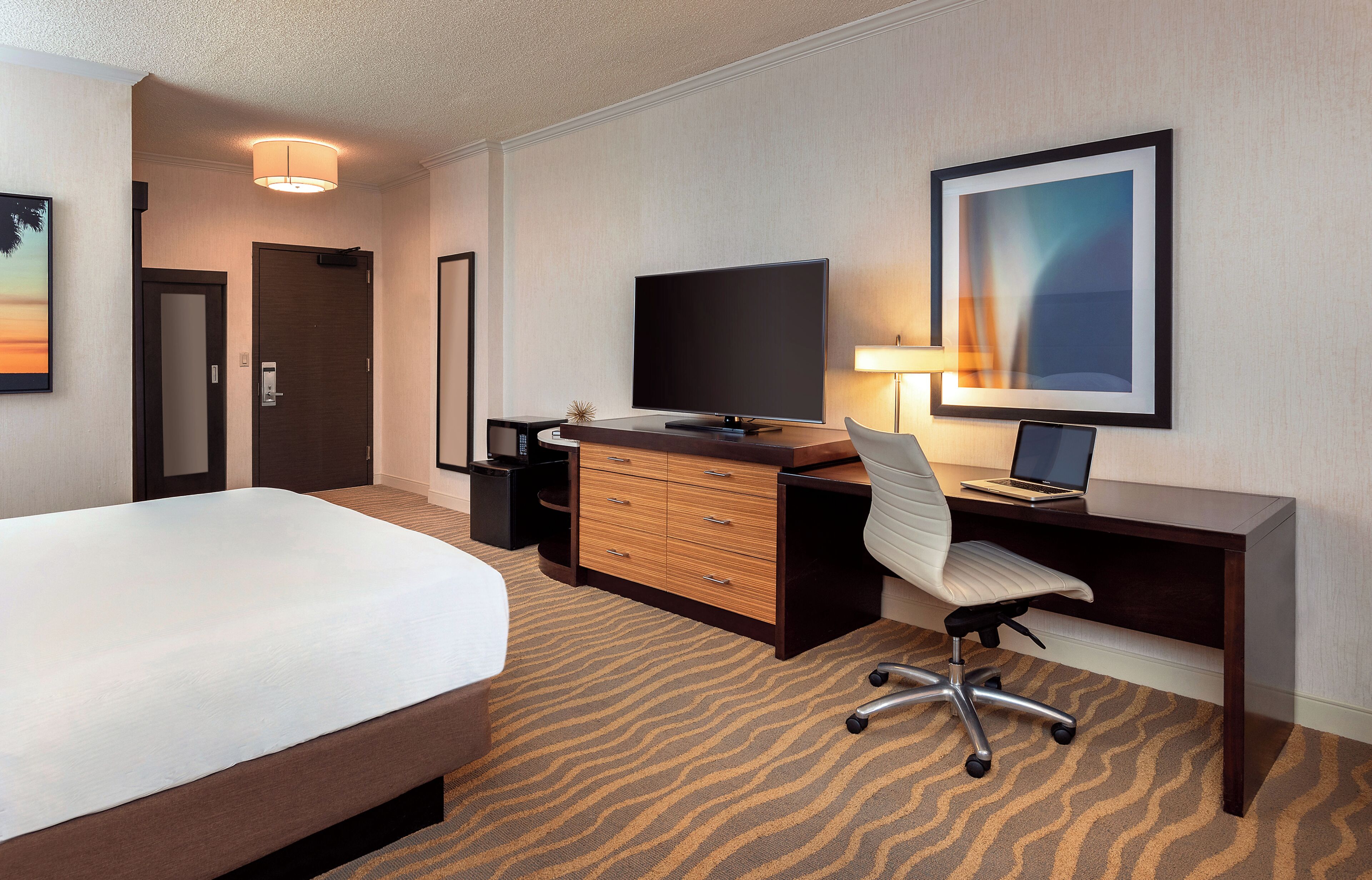 Una habitación de hotel con cama, escritorio con computadora, televisor, silla, lámpara y un cuadro en la pared.